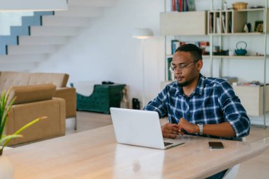 Modern bir oturma odasındaki masada oturan gözlüklü Afrikalı Amerikalı etkileyiciler iş görüşmesi, arkadaşlarla sohbet ve eğlence için dizüstü bilgisayar kullanıyorlar.