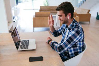 Modern bir oturma odasındaki masada oturan bir serbest çalışan, kulaklıklarıyla iş görüşmesi için bir laptop kullanan, arkadaşlarıyla sohbet eden ve eğlenen biri..