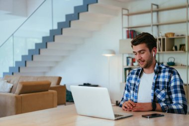 Modern bir oturma odasındaki masada oturan bir iş adamı, kulaklıklarıyla iş videosu sohbeti, arkadaşlarla sohbet ve eğlence için dizüstü bilgisayar kullanıyor.