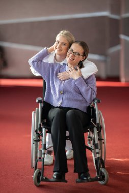 Gülümseyen bir hemşire, tekerlekli sandalyedeki yaşlı bir hastayı kucaklar ve sağlık hizmetlerinde şefkat ve bakım gösterir..