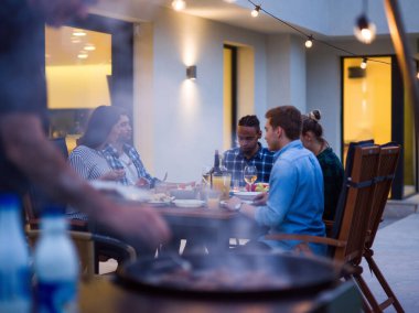 Bir grup genç insan akşamları modern bir evin terasında yemek yiyorlar. Dostlar ve aile için eğlence. Tatil kutlamaları, barbekülü düğünler..