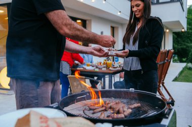 Bir grup arkadaş ve aile akşamları terasta büyük, modern bir evin önünde barbekü yapıyorlar.