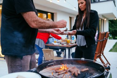 Bir grup arkadaş ve aile akşamları terasta büyük, modern bir evin önünde barbekü yapıyorlar.