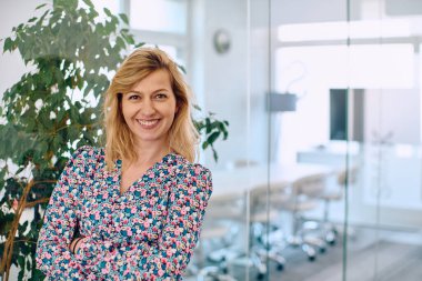 Renkli elbiseli gülümseyen bir iş kadını şirket koridorunda zarafetle duruyor. İş yerinde profesyonellik ve canlılığı somutlaştırıyor..