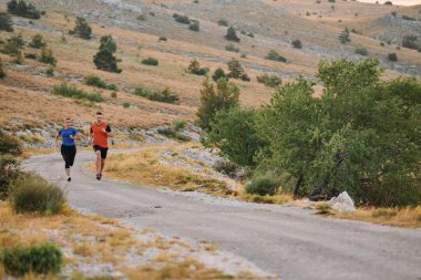 Spor kıyafetleri giymiş bir çift sabahın erken saatlerinde spor yaparken manzaralı bir yolda koşuyor, temiz havanın tadını çıkarıyor ve sağlıklı bir yaşam tarzı sürdürüyorlar..