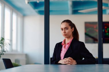 Pembe gömlekli başarılı bir bayan lider modern cam bir ofiste oturmuş kararlı bir gülümsemeyle bekliyor.