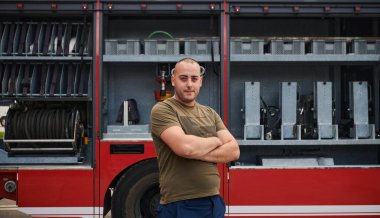 Kendine güvenen itfaiyeci, çapraz kollarla ayakta duruyor, dayanıklılık ve hazırlık yayıyor, modern bir itfaiye aracının yanında acil durumlara cevap vermeye hazır, ateşin kahramanlığını ve gücünü gösteriyor.
