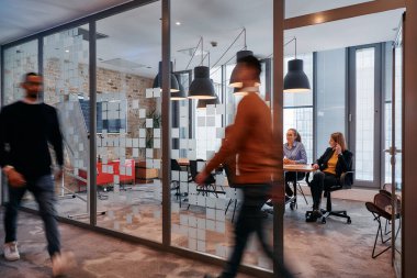 İş ortamının hareketli koşuşturmasında, bir grup genç iş adamı ofislerinin yanındaki koridorda yürürken, meslektaşları içeride iş birliği yaparak,