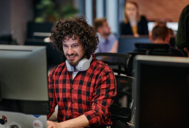 Afro saç stili ve ekose gömlekli bir programcı, modern ofis ortamındaki bir bilgisayarda özenle kodluyor.