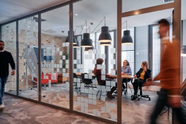 İş ortamının hareketli koşuşturmasında, bir grup genç iş adamı ofislerinin yanındaki koridorda yürürken, meslektaşları içeride iş birliği yaparak,