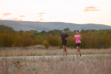 Spor kıyafetleri giymiş bir çift sabahın erken saatlerinde spor yaparken manzaralı bir yolda koşuyor, temiz havanın tadını çıkarıyor ve sağlıklı bir yaşam tarzı sürdürüyorlar.