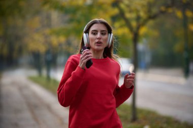 Sağlıklı yaşam tarzı, sabah koşusu. Genç ve güzel bir kadın sonbahar parkında koşuyor ve akıllı telefonuyla kulaklıkla müzik dinliyor. Kırmızı bir spor gömlek giyiyor.. 