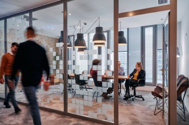 İş ortamının hareketli koşuşturmasında, bir grup genç iş adamı ofislerinin yanındaki koridorda yürürken, meslektaşları içeride iş birliği yaparak,