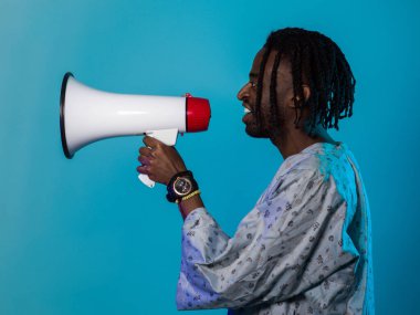 Afro-Amerikan erkek geleneksel kıyafetler giyer, çarpıcı bir mavi arkaplan karşısında megafonu tutkulu bir şekilde kullanır, sosyal adalet arayışındaki ses ve kültürel gücünü sembolize eder.