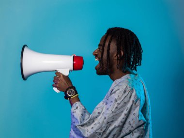 Afro-Amerikan erkek geleneksel kıyafetler giyer, çarpıcı bir mavi arkaplan karşısında megafonu tutkulu bir şekilde kullanır, sosyal adalet arayışındaki ses ve kültürel gücünü sembolize eder.