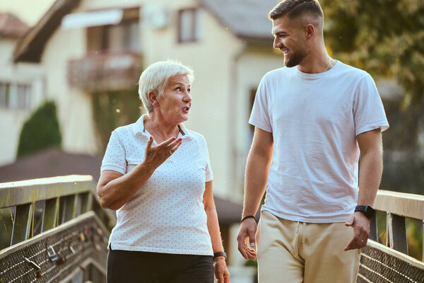 A handsome man and an older woman share a serene walk in nature, crossing a beautiful bridge against the backdrop of a stunning sunset, embodying the concept of a healthy and vibrant intergenerational