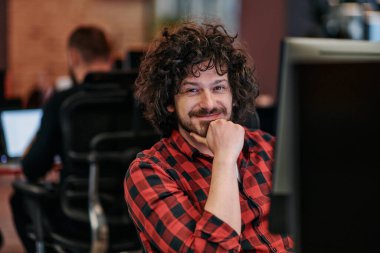 Afro saç stili ve ekose gömlekli bir programcı, modern ofis ortamındaki bir bilgisayarda özenle kodluyor.