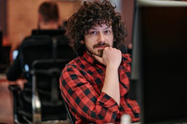 Afro saç stili ve ekose gömlekli bir programcı, modern ofis ortamındaki bir bilgisayarda özenle kodluyor.