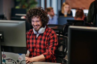 Afro saç stili ve ekose gömlekli bir programcı, modern ofis ortamındaki bir bilgisayarda özenle kodluyor.