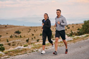 Spor kıyafetleri giymiş bir çift sabahın erken saatlerinde spor yaparken manzaralı bir yolda koşuyor, temiz havanın tadını çıkarıyor ve sağlıklı bir yaşam tarzı sürdürüyorlar..
