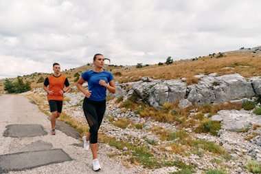 Spor kıyafetleri giymiş bir çift sabahın erken saatlerinde spor yaparken manzaralı bir yolda koşuyor, temiz havanın tadını çıkarıyor ve sağlıklı bir yaşam tarzı sürdürüyorlar..