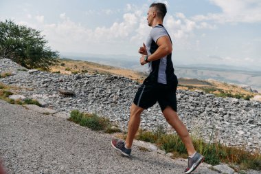 Kaslı bir erkek sporcu gün doğumunda engebeli bir dağ yolunda koşar, nefes kesen kayalık manzaralar ve doğal güzelliklerle çevrilidir.. 