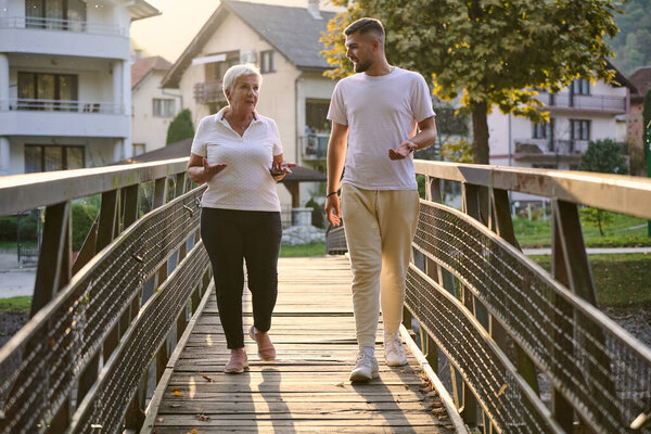 A handsome man and an older woman share a serene walk in nature, crossing a beautiful bridge against the backdrop of a stunning sunset, embodying the concept of a healthy and vibrant intergenerational