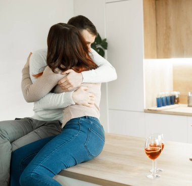 Birbirine aşık genç bir çift modern bir mutfakta kırmızı şarap içip sarılıyorlar. Sevgi ve şefkat kavramı.