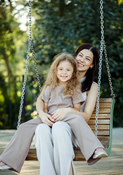 Mom and daughter swing on a swing. Caucasian woman and little girl have fun on the playground. Happy family. Summer day.