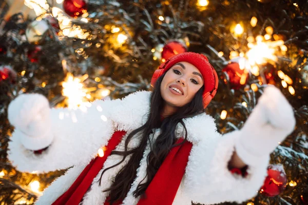 迷人的年轻女子头戴红帽子 头戴耳机 在圣诞树旁的圣诞市场上手握火花 快乐时光 — 图库照片