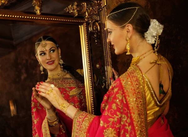 Female model Hindu Bride in saree, wearing gold and jasmine flower garlands in the hair looks in the mirror and touches his reflection, back view.