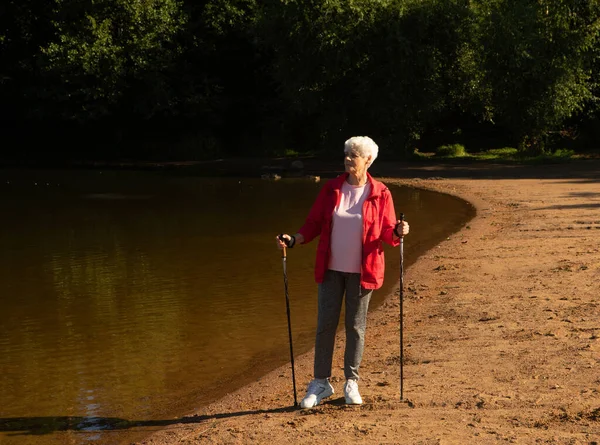 头发灰白的女人带着跟踪棒在湖边散步 生活方式的概念 — 图库照片