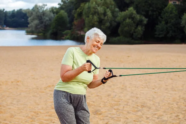 长发的老妇人穿着运动服在海滩上做运动 用的是阻力橡皮筋 健康的生活方式 积极的退休生活和运动时间 — 图库照片
