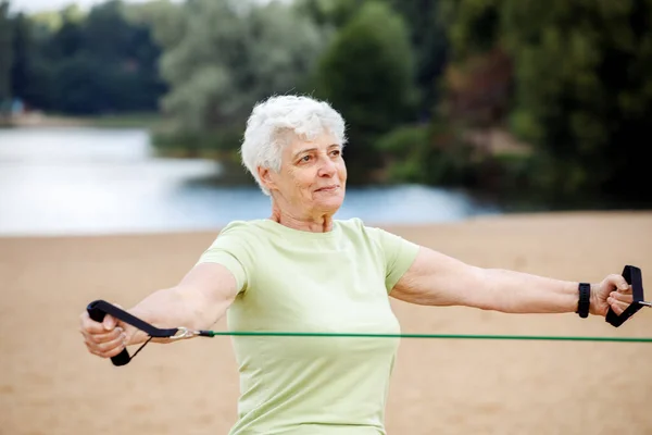 长发的老年妇女穿着运动服在海滩上做运动 用的是阻力橡皮筋 健康的生活方式 积极的退休生活和运动时间 — 图库照片