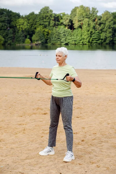 长发的老年妇女穿着运动服在海滩上做运动 用的是阻力橡皮筋 健康的生活方式 积极的退休生活和运动时间 — 图库照片