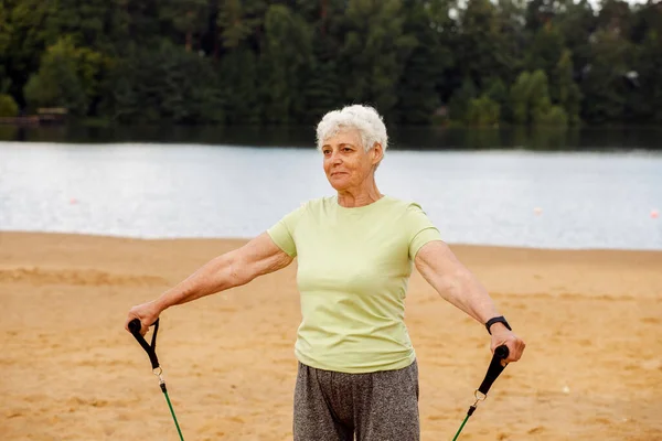 长发的老妇人穿着运动服在海滩上做运动 用的是阻力橡皮筋 健康的生活方式 积极的退休生活和运动时间 — 图库照片