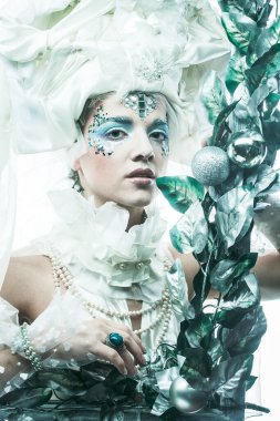 Parti, Noel ve insanlar: Noel karnavalı için kostüm giymiş güzel bir kadın Noel süslemeleri ve gümüş yapraklar çerçevesinde poz veriyor. Yaratıcı makyaj, saç stili.