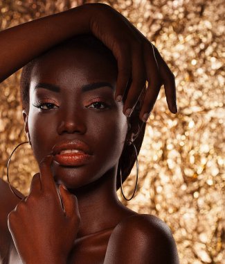 Koyu tenli, genç ve güzel bir kadın, altın bir arka planda şehvetle poz veriyor. Parlak makyajlı kusursuz bir ten..