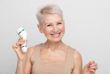 Beautiful 60s woman holding a massager in her hand. Spa technique skincare ads. Portrait over grey background. clipart