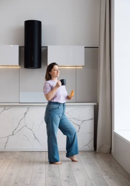 Kapalı alanda, pencerenin yanında kahve içen, kıvrımlı, esmer bir kadın resmi. Neşeli tombul kız evde dinleniyor, kruvasanla fincandan çay içiyor..