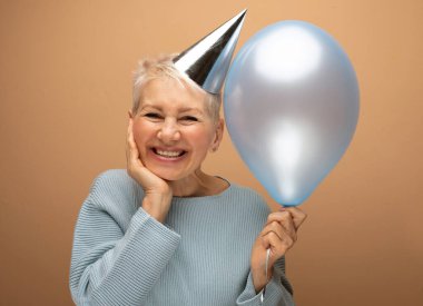 Güzel görünümlü, çekici, neşeli, neşeli, neşeli, gri saçlı, elinde mavi balonla gezen beyaz bej arka planda izole edilmiş bir kadının portresi.