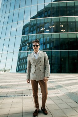 İş dünyası, insanlar ve başarı kavramı: Şaşılası kıyafetli yakışıklı genç iş adamı ve güneş gözlüğü modern ofis yakınlarında yaz mevsiminde yürüyor