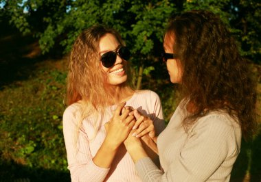Büyük anne ve yetişkin kızı bir yaz parkında el ele tutuşuyorlar, güneş gözlüğü takan mutlu kadınlar.