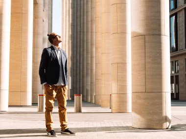 Yaşam tarzı, iş ve insan konsepti: Modern büyük bir binanın sütunlarının yanında sıradan giyinmiş genç zarif yakışıklı adam. Yaz günü.