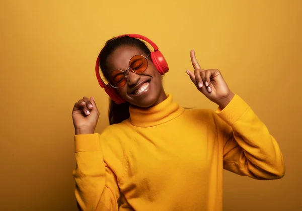 People, music, emotions concept. Delighted carefree afro american female with dances in rhythm of melody, listens loud song in headphones, holds smartphone has fun.