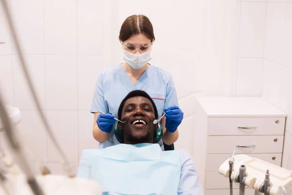 专业的女牙医检查男人的牙齿 现代门诊部接受牙科治疗的黑人男性患者的画像 — 图库照片