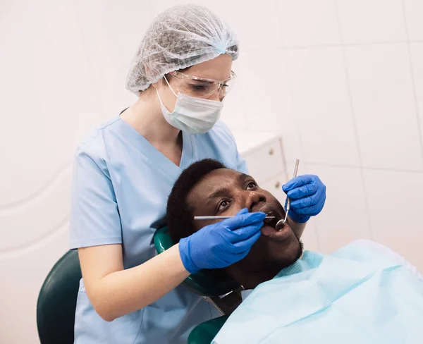 在现代门诊部接受牙医治疗的黑人男性患者的肖像 坐在白色橱柜的椅子上 女性专业口腔科医生 使用牙钻工具为年轻人提供服务 — 图库照片