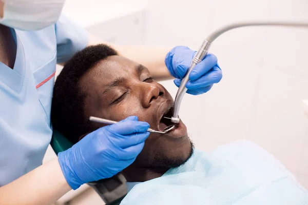 在现代门诊部接受牙医治疗的黑人男性患者的肖像 坐在白色橱柜的椅子上 女性专业口腔科医生 使用牙钻工具为年轻人提供服务 — 图库照片