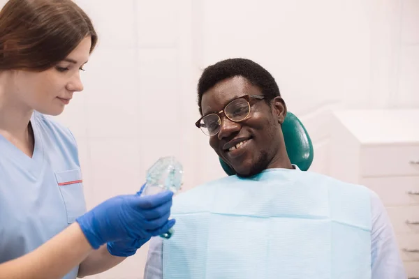 年轻的女牙医在现代牙科诊所做检查时 给一个黑人黑人男子展示假牙 展示了一个牙齿的例子 — 图库照片