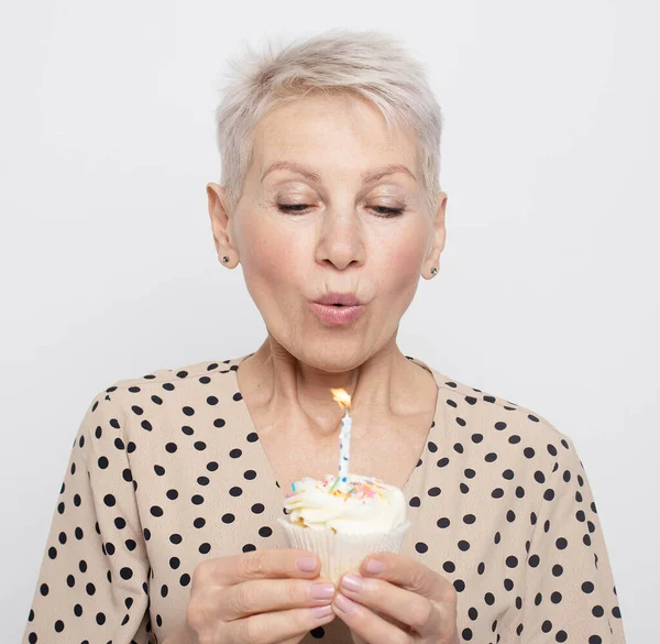 Portrait of elderly woman holding birthday cupcake making wish with eyes closed and blowing out candle celebrating anniversary. Happy mature lady with party cake.